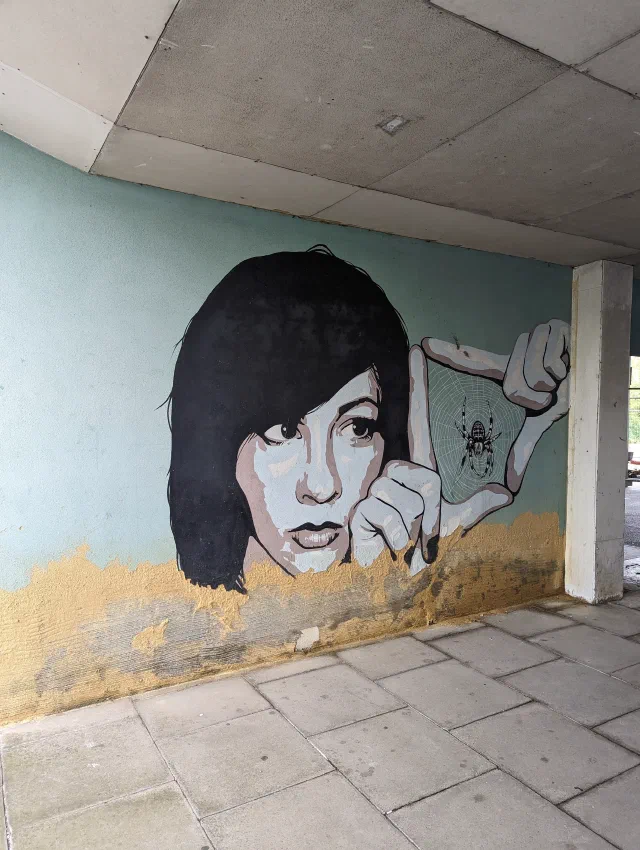Black and white painting of a woman with a dark bob, holding a beetle between her fingers, on a pale blue wall under a concrete overhang.