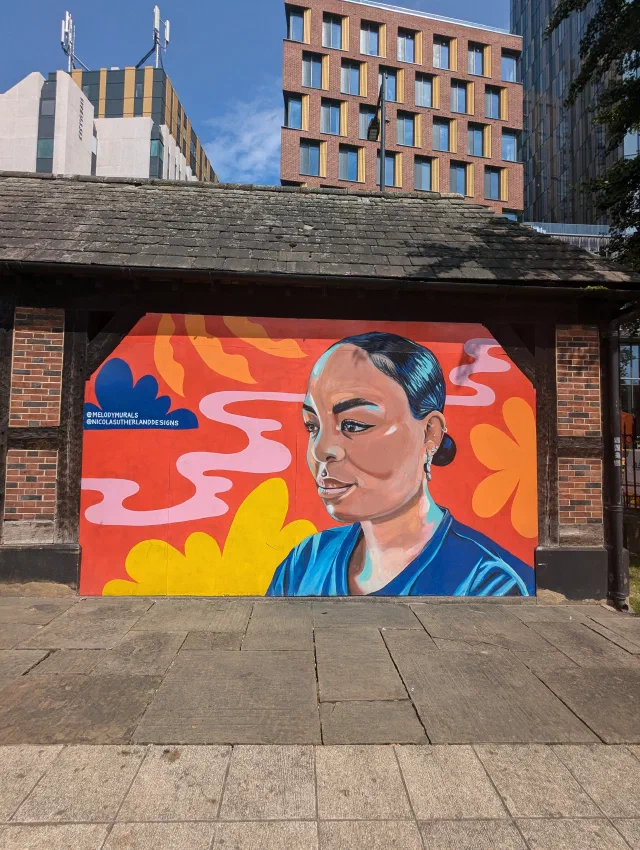 Mural of a woman in a blue top with her hair in a bun, in front of bright orange, pink and yellow swirls, on the end of a small brick building.