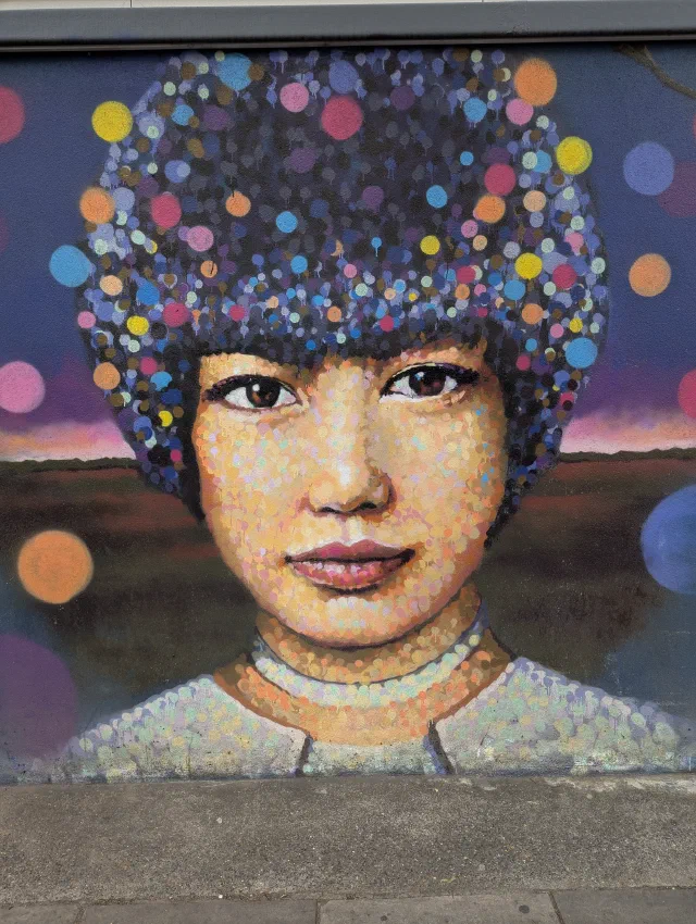 A woman's face painted in dots of colour, her hair a cloud of bright spots against a purple sky.