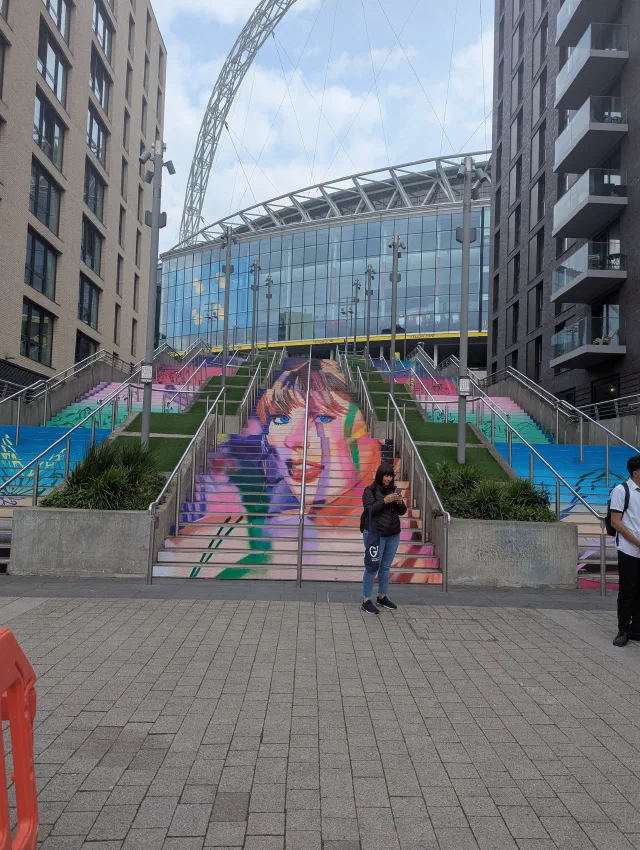 A woman's face painted across a wide flight of steps, with Wembley Stadium's arch behind.