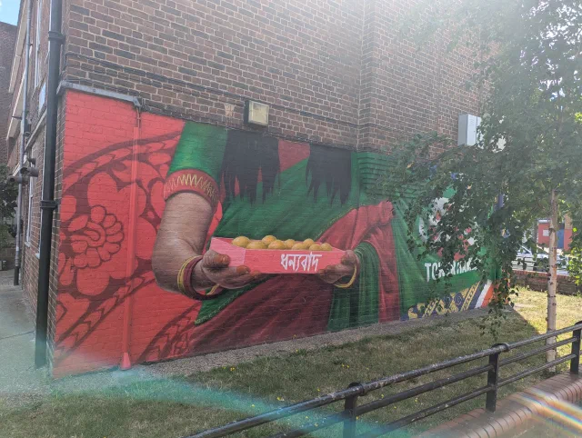 Mural of a hand with a gold bangle and a red and green sari, holding out a tray of sweets labelled "thank you" in Bengali, on a brick wall behind railings.