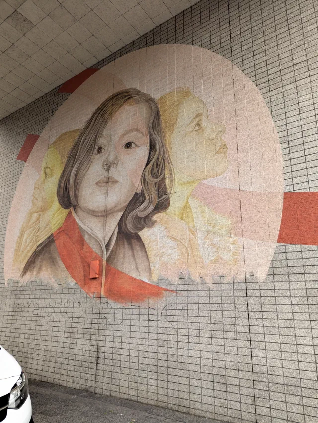 Two women painted in soft pinks and oranges, one face over the other, on a white tiled wall.