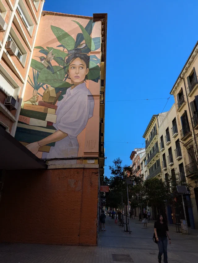 A woman in a white top among potted plants, painted high on the end wall of a tall building over a narrow street.