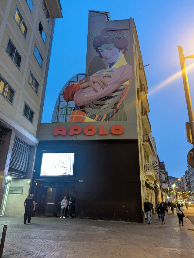A woman with her arms folded, painted high above a theatre sign reading APOLO, at dusk.