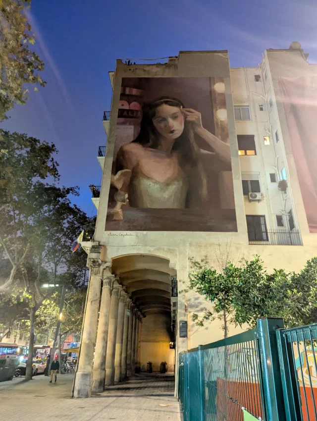 A woman in a pale dress touching her hair, painted as a framed portrait high on a building above an arched arcade, at dusk.
