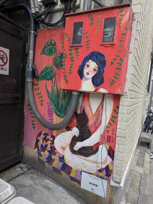 A woman with blue hair holding a black cat, painted on a red wall and electrical box in a narrow alley.