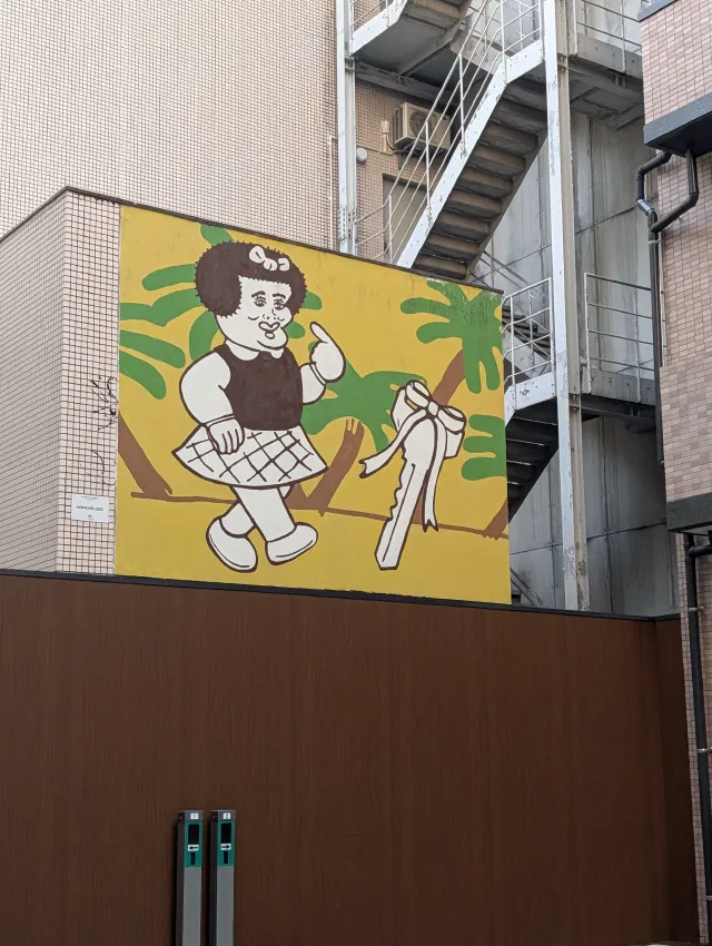 Cartoon of a girl in a checked skirt, in black and white on a yellow wall with palm leaves.