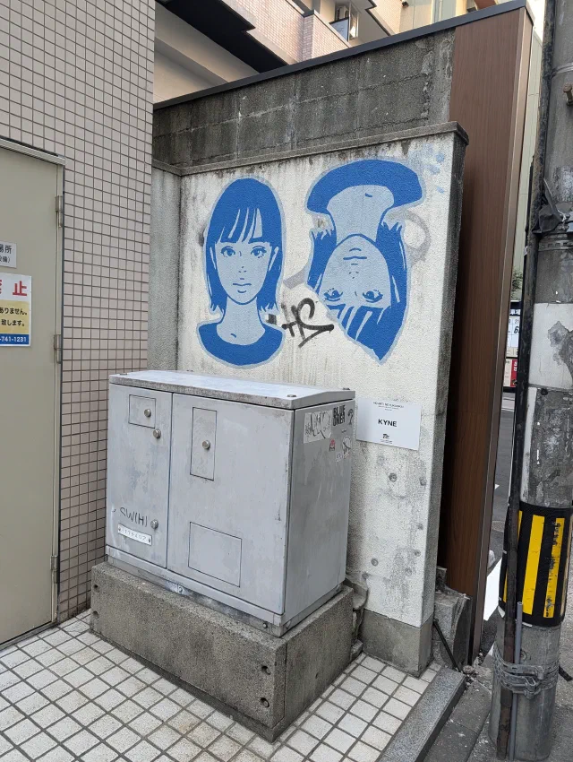 Two blue-haired faces, one upside down, painted on a white panel above a grey electrical cabinet.