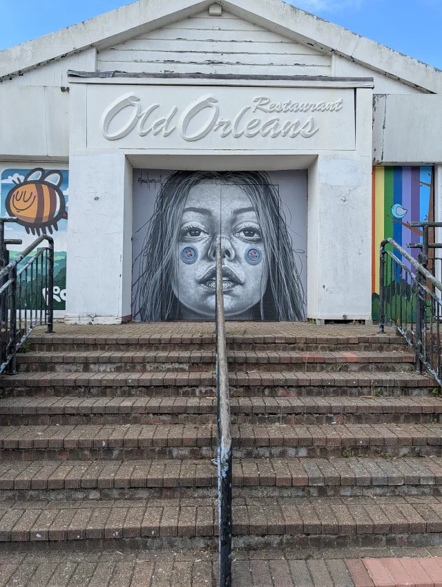 Grey portrait of a woman with blue circles on her cheeks, filling the doorway of an old restaurant at the top of a flight of steps.