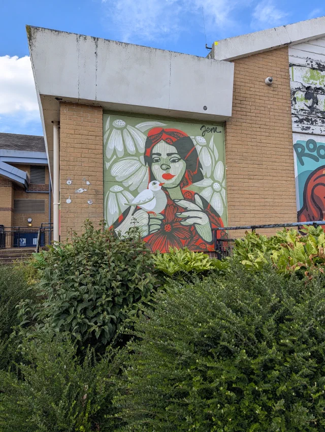 A woman in red holding a white bird, painted on a panel above a row of bushes.