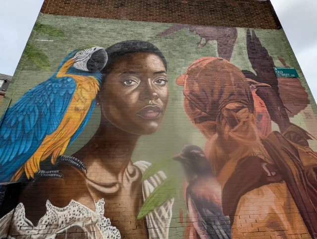 Two women painted on a brick end wall, one with a blue and yellow macaw on her shoulder.
