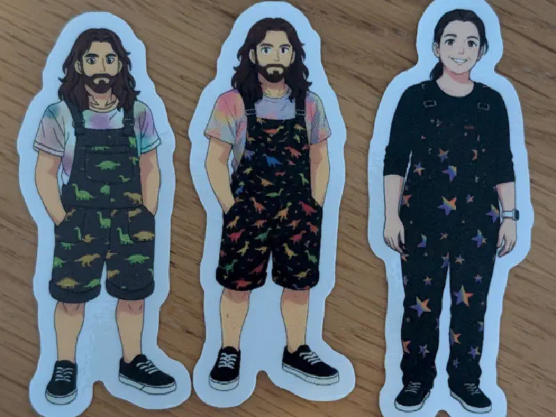 Three die-cut character stickers on a wooden table: two of me in dinosaur dungarees and one of my partner