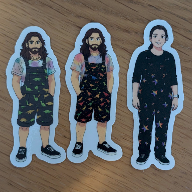 Three die-cut character stickers on a wooden table: two versions of me in dinosaur-print dungarees and one of my partner in dark star-pattern overalls.