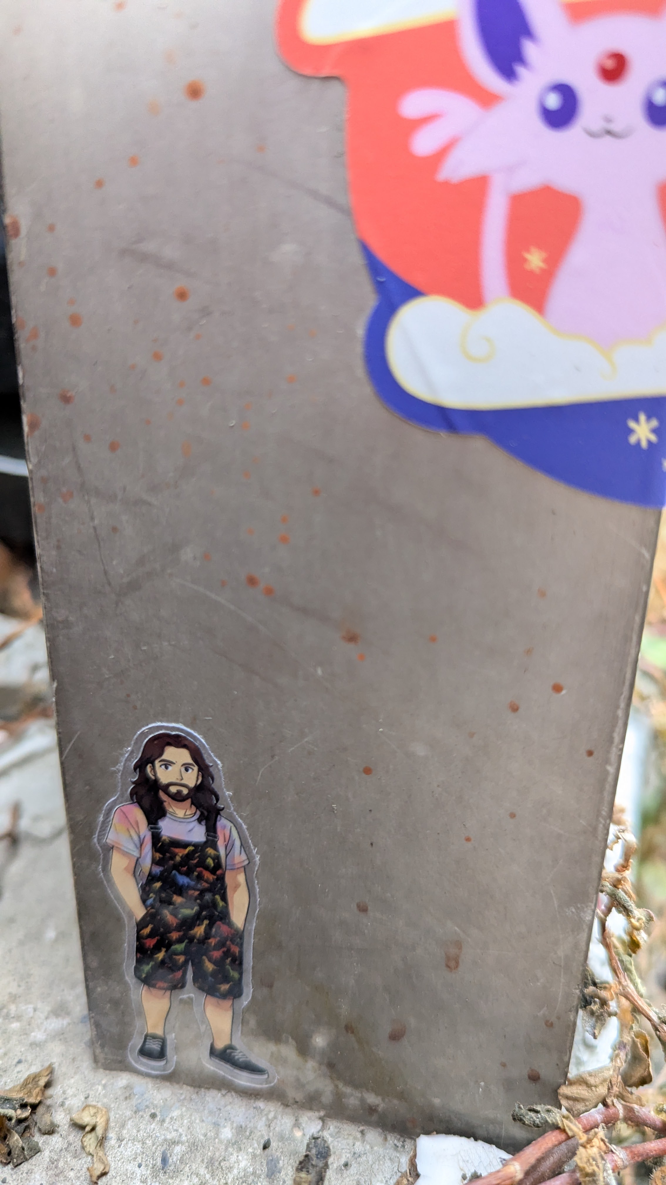 Small die-cut sticker of a bearded person (me) with long dark hair, a pastel striped T-shirt and dark dinosaur-print dungarees stuck to a weathered metal post outdoors.