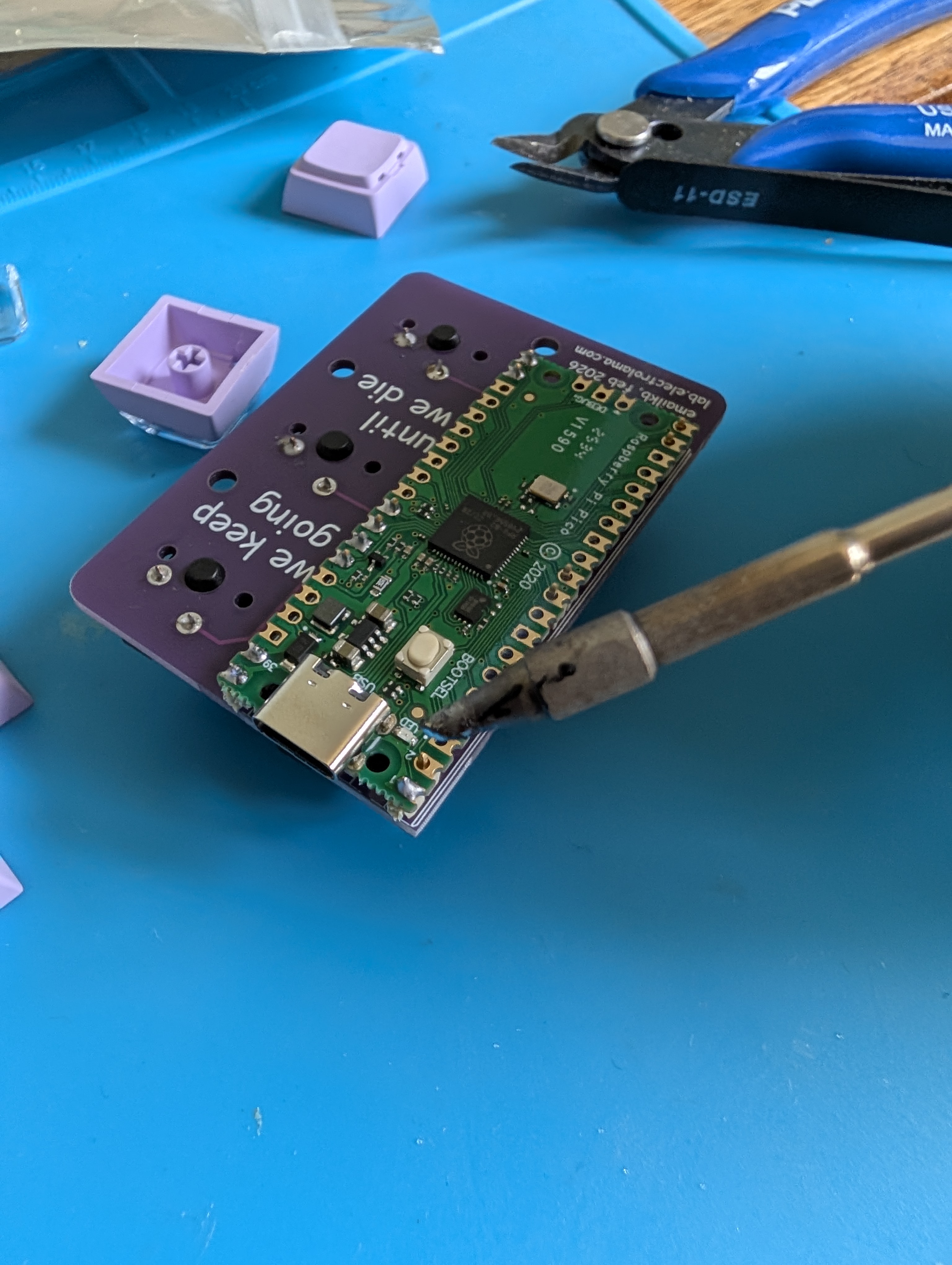 The emailkb PCB held at an angle with pliers on a blue surface, a soldering iron tip touching the pads to attach the Raspberry Pi Pico to the back of the board.