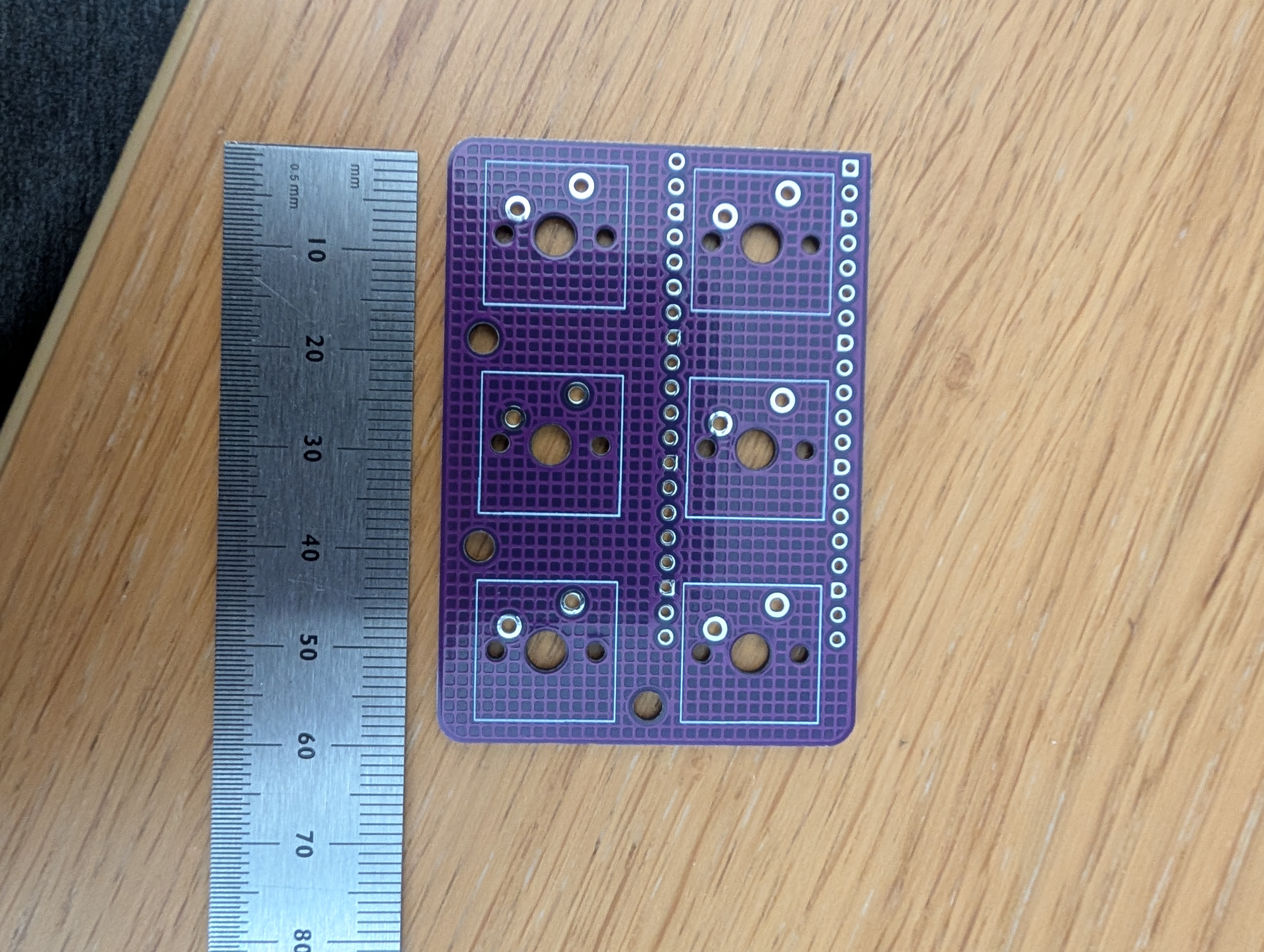 Front face of the purple emailkb PCB on a wooden table next to a metric ruler, showing the six MX switch footprints arranged in a two-by-three grid with through-holes down the right edge for the Pico.