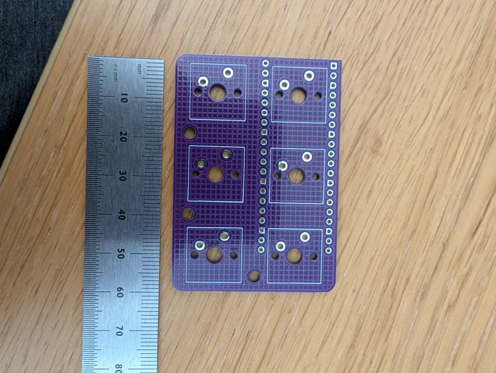 Front face of the purple emailkb PCB on a wooden table next to a metric ruler, showing the six MX switch footprints arranged in a two-by-three grid with through-holes down the right edge for the Pico.