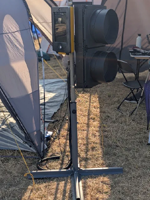 The beg button in daylight: a yellow push-button unit with a screen, beside a black two-aspect Walk/Stop light, on a pole with a cross-shaped stand on dry grass between tents.