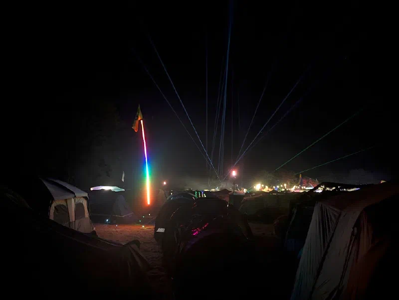 Tents in a dark field at night, with laser beams fanning up into the sky from the stage area and a rainbow LED pole flying a flag.