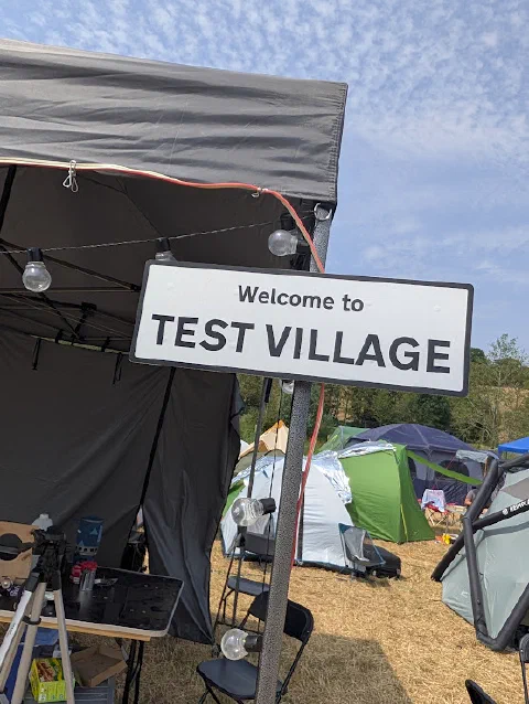 A white road-style sign reading Welcome to TEST VILLAGE on a pole beside a grey gazebo strung with festoon bulbs, with tents and a blue sky behind.