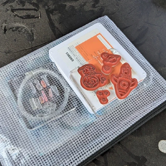 Red rubber stamps of a rocket, a ringed planet, a cartoon figure and some stars on a clear block, next to a ring of clear letter stamps, in a mesh zip bag.