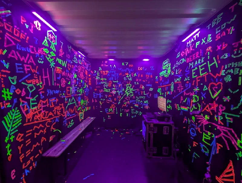 A room lit by UV tubes, its black walls covered floor to ceiling in neon tape graffiti (names, hearts, EMF, I LOVE TRAINS), with a bench along one side and a screen on a flight case at the end.