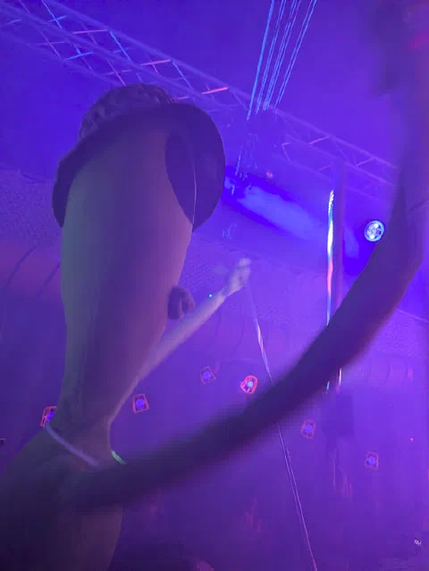 A mannequin in a bucket hat with one arm raised, hanging under the lighting rig in purple light.
