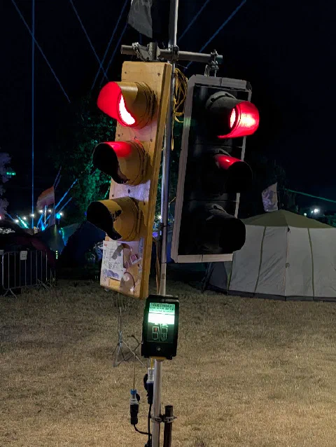 Another installation at night: two sets of traffic lights showing red on a pole, one in a weathered yellow housing, above a pedestrian push-button box with a glowing green display, with lasers in the sky behind.