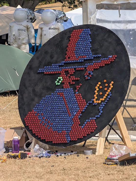 A large round black board standing in a field, covered in red, blue and orange bottle caps that make a pixel-art wizard in a tall hat, with two astronaut figures behind it.