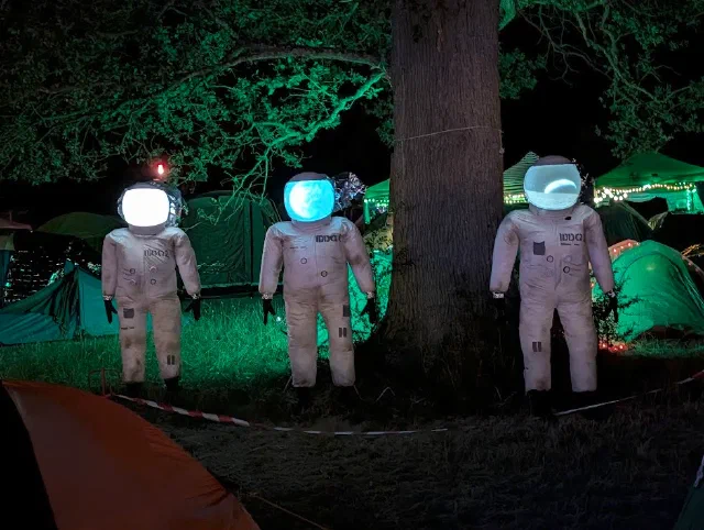 Three astronaut figures standing under a tree among tents at night, their helmet visors glowing as screens.
