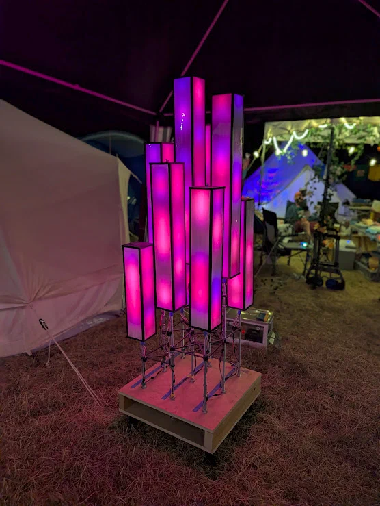 A cluster of tall translucent columns of different heights on a wooden base, glowing pink and purple under a tent canopy at night.