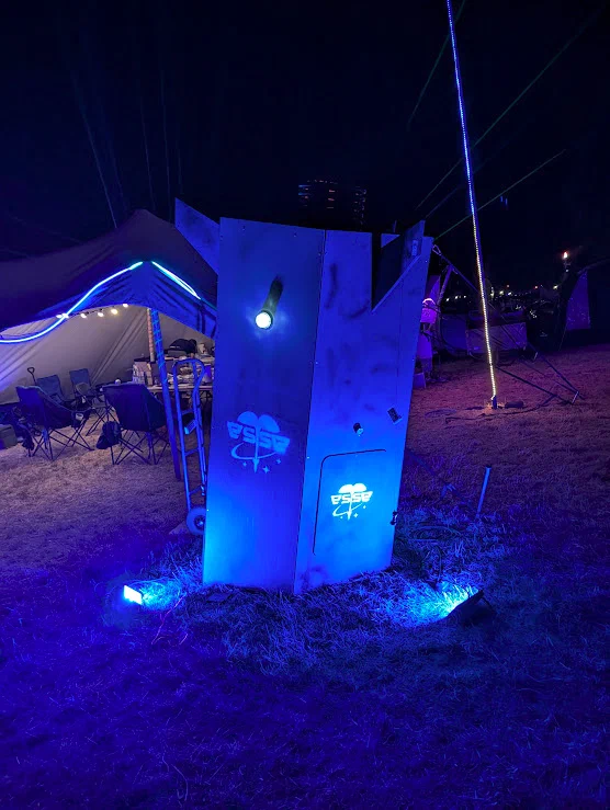 A tall angular wooden structure lit blue at night, with a glowing space-agency style logo, a small spotlight and a door panel, in a field beside a gazebo.