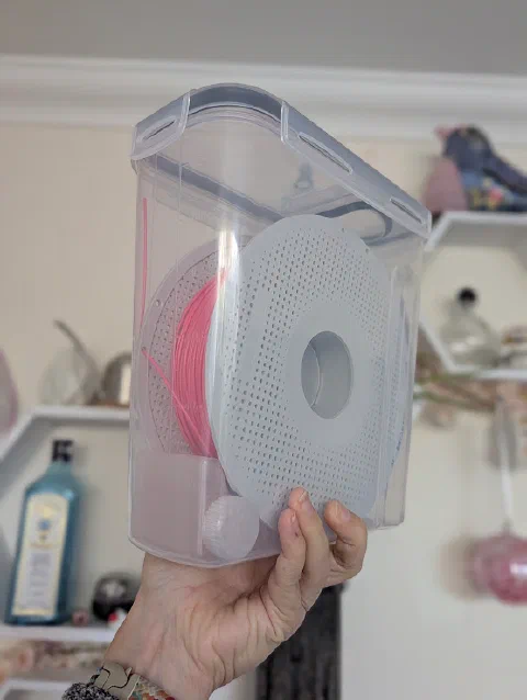 A hand holding a clear plastic cereal container with a clip-down lid. Inside is a spool of pink filament standing on its edge, with a translucent printed desiccant holder in the bottom corner.