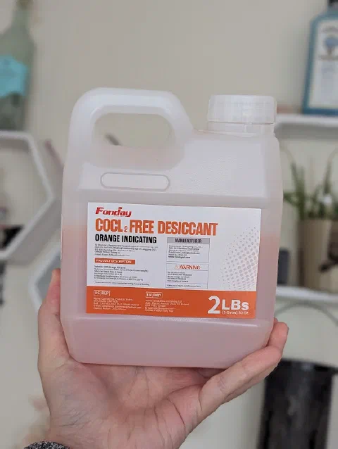 A hand holding a translucent plastic jug of orange silica gel beads, labelled Fonday CoCl2 Free Desiccant, Orange Indicating, 2 LBs.