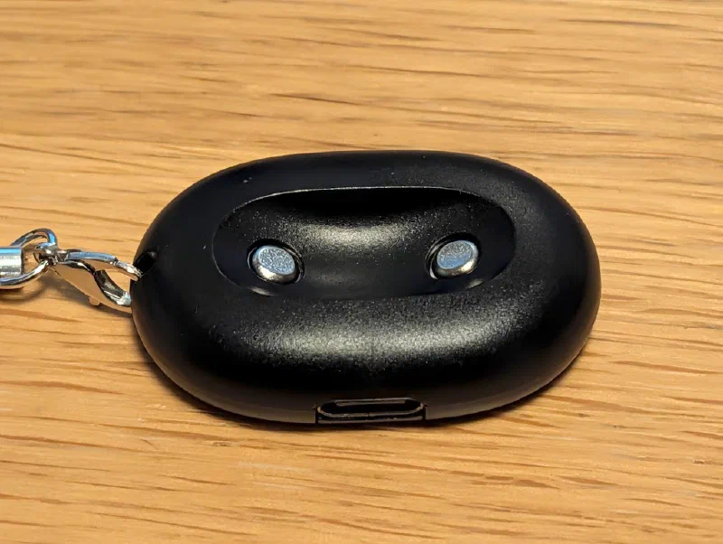 A small black oval charging puck on a wooden table, with a shallow curved recess on top holding two round metal charging pins, a USB-C port on the front edge, and a silver lobster clasp and keyring clipped through a hole on the left end.