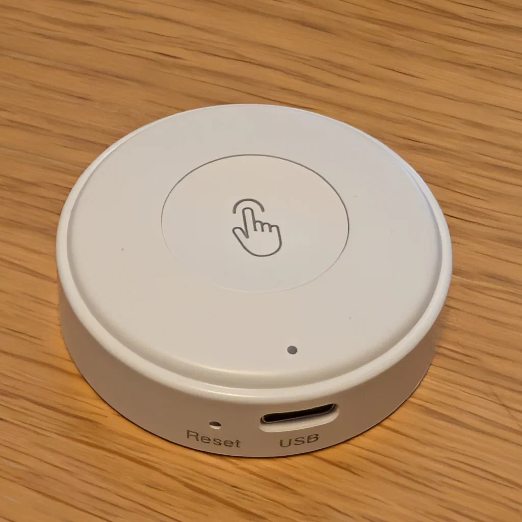 A round white puck-shaped button on a wooden table, with a recessed circular pad printed with a grey pointing-finger icon, a small LED dot on the side, and a reset pinhole and USB-C port labelled Reset and USB on the edge.