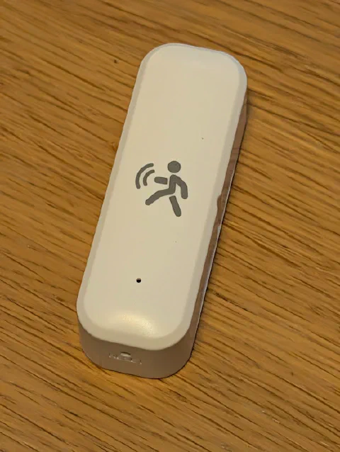 A small white pill-shaped sensor on a wooden table, with a grey icon of a walking figure and radar waves on its face, a pinhole near the bottom and a small hole in the end.
