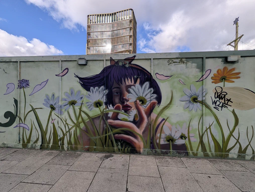 A woman with a dark purple bob peering through a clump of white daisies and long grass, painted on pale green hoarding under a blue sky.