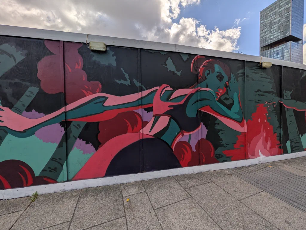 The same mural from further back: the woman stretched along the hoarding among birch trees and red clouds.