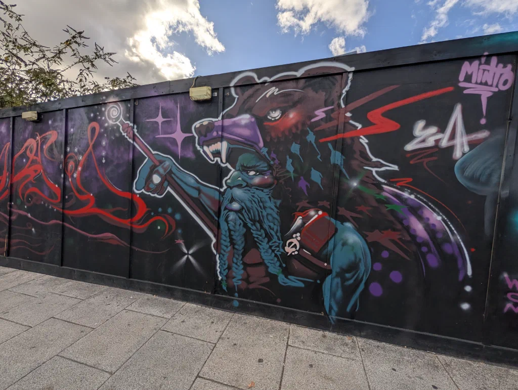 A blue bearded wizard with a staff, in front of a snarling bear, with stars and red lightning on dark purple hoarding.