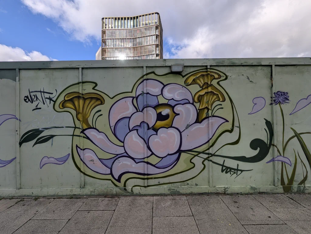 A large pale lilac flower with an eye at its centre, flanked by two golden mushrooms, painted on pale green hoarding with a tall building behind.