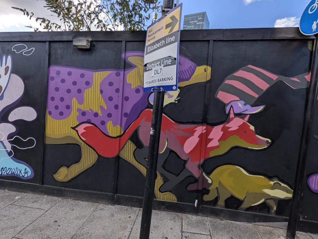 A red fox in a striped hat and a yellow badger running past a big purple and yellow animal, behind a rail replacement bus sign.