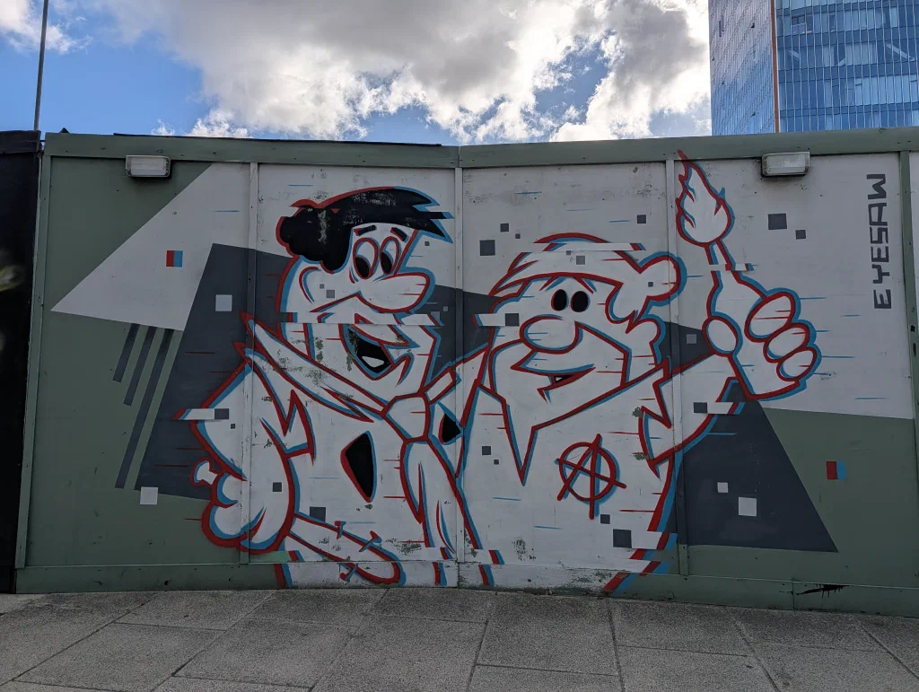 Fred Flintstone and Barney Rubble painted in white with red and blue outlines, as if glitching, on grey and green hoarding.
