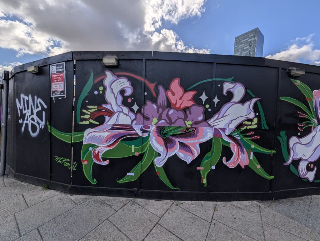 Large pink and white lilies with green leaves painted along curving black hoarding, next to a security sign.
