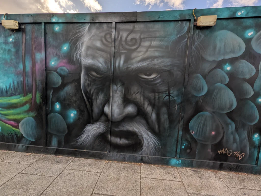 A black and white portrait of an old man with a drooping moustache and a spiral painted on his forehead, among glowing blue mushrooms.