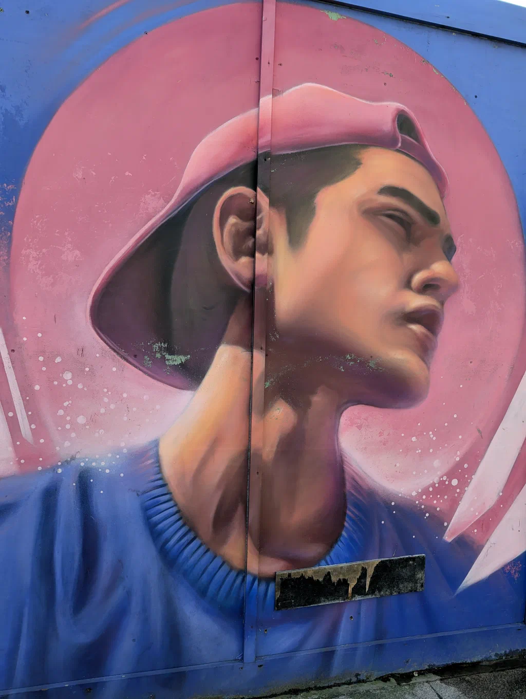 A young man in a backwards pink cap and blue jumper, looking up, inside a pink circle on blue hoarding.