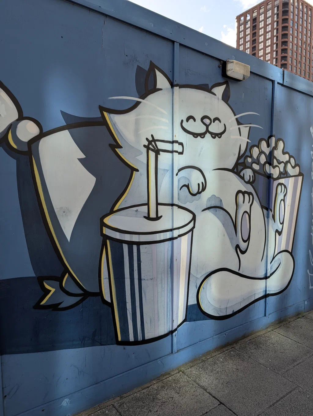 A fat white cartoon cat sipping from a giant cup with a straw, next to a bucket of popcorn, on blue hoarding.