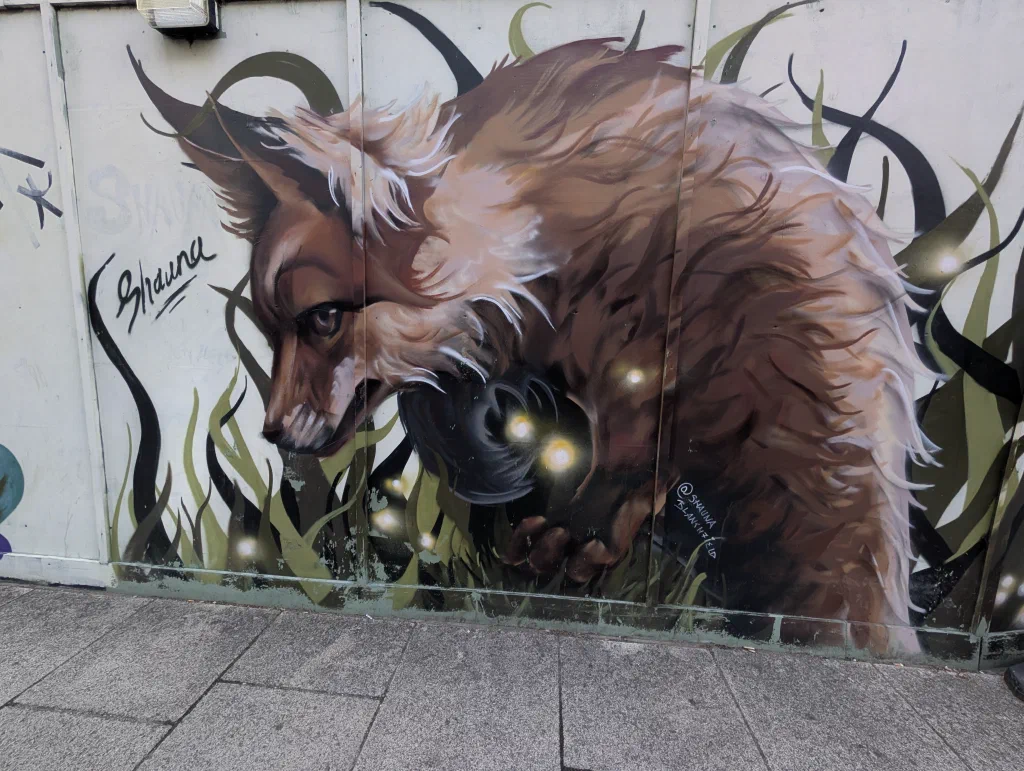 A red fox with a shaggy coat sniffing at the ground among dark grass and glowing fireflies, painted on white hoarding.