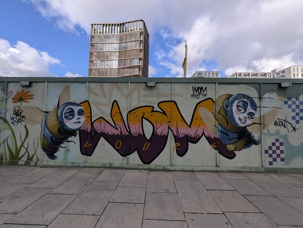 The letters WOM in orange, pink and purple, with two bees with pale cartoon faces flying either side.