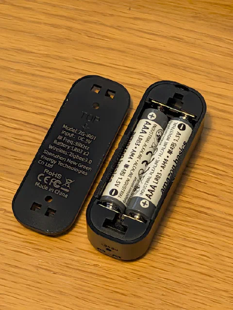The ZG-IR01 with its back cover off, showing two Amazon Basics AAA batteries. The cover's label reads Model ZG-IR01, Input DC 3V, IR Freq 38KHz, Battery LR03 x2, Wireless Zigbee3.0, Shenzhen New Green Energy Technologies Co Ltd.